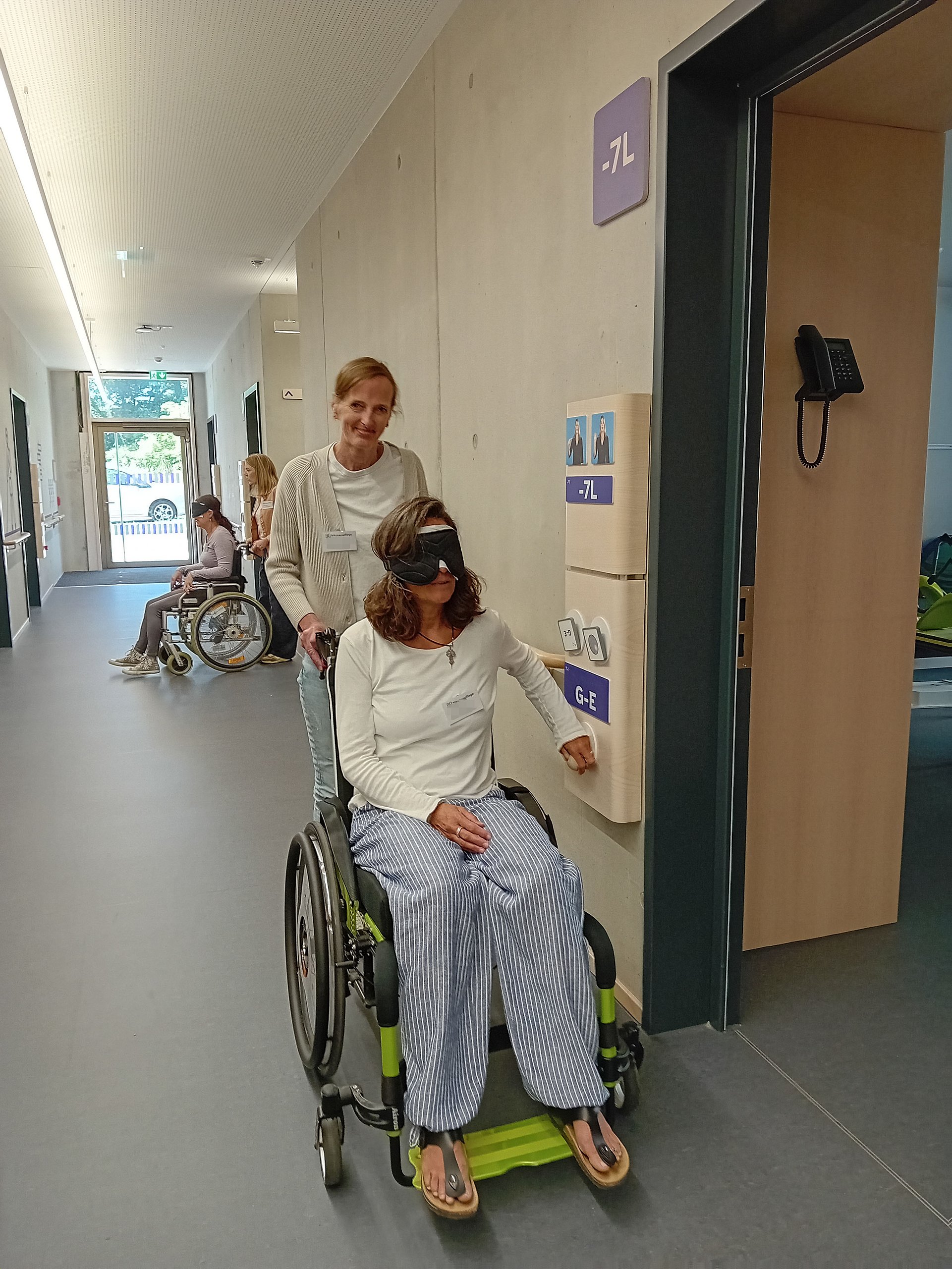Eine Frau in einem Rollstuhl mit Augenbinde in einem Flur. Hinter dem Rollstuhl steht eine Person