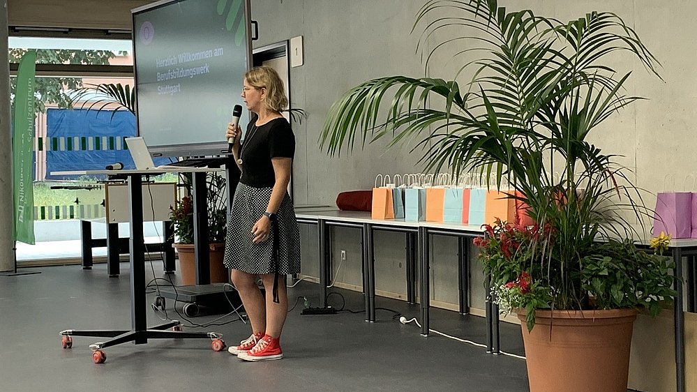 Eine Frau mit schwarz-weißem Rock und schwarzer Bluse steht vor einem Smartboard an einem Stehtisch. Sie spricht in ein Mikrofon. Hinter ihr steht ein Tisch mit vielen kleinen bunten Tüten darauf.