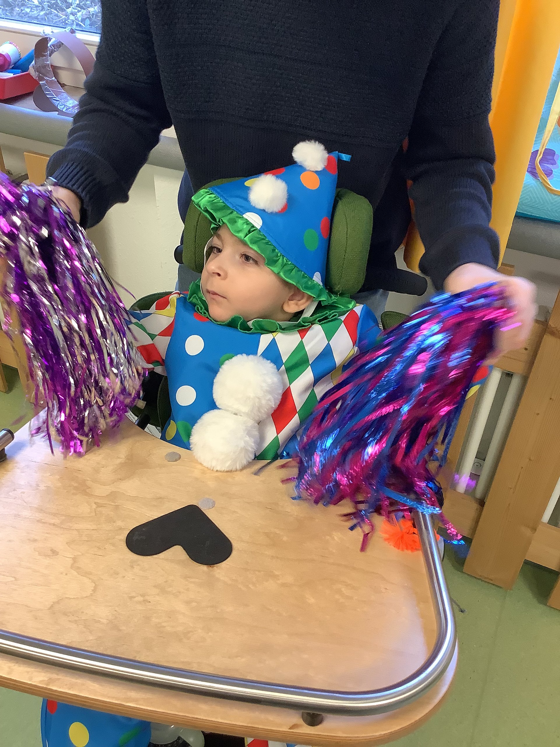 Ein kleiner Junge sitzt in einem Rollstuhl. Er ist als Clown verkleidet. Hinter ihm steht eine Betreuerin und schwenkt mit ihm gemeinsam glitzernde Pompons