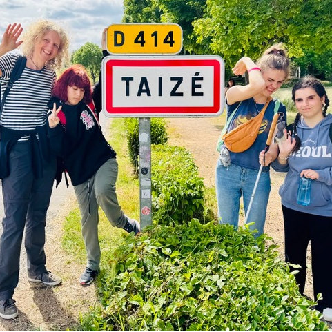 Eine Frau steht mit weiteren drei jungen Frauen auf der Straße neben einem Straßenschild. Auf diesem ist der Name einer französischen Stadt zu lesen: Taisé