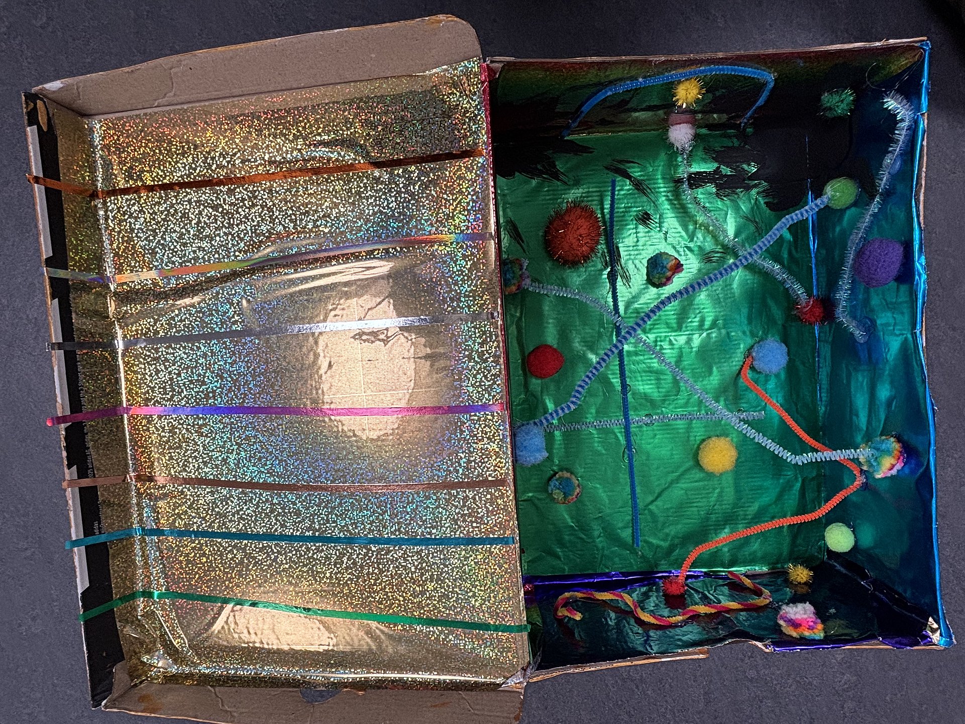 Ein aufgeklappter bunt gestalteter Pappkarton. Links mit goldenem Glitzer und Bändern, rechts in Grün und Dekoelemente