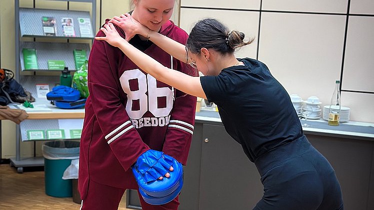 Zwei Jugendliche stehen sich gegenüber, die eine hält zwei Standboxsäcke vor sich, die andere hält sich mit ihren Händen an der Schulter der anderen fest und tritt gegen die Boxsäcke.