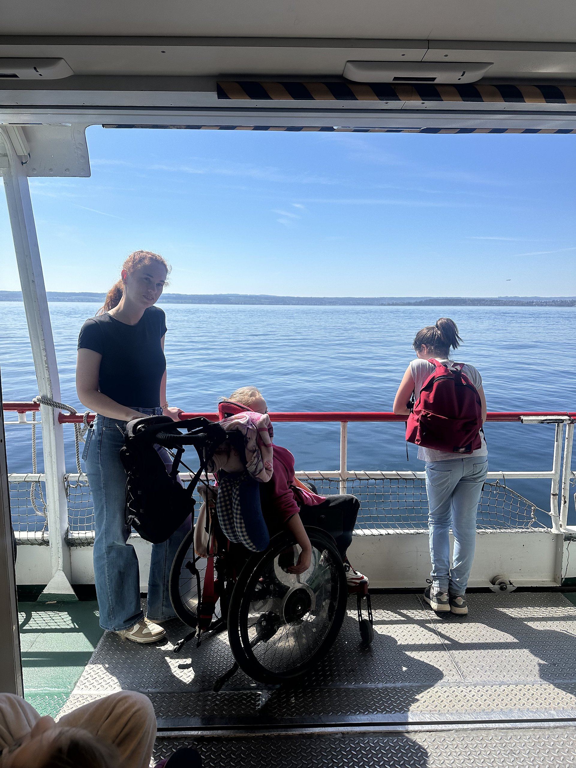 Eine junge Frau steht an deck eines Schiffes. Es fährt über den Bodensee und die Sonne scheint. Ihre Hände liegen auf dem Lenker eines Rollstuhles, in dem eine Klientin sitzt und auf den See schaut. Rechts neben ihnen steht eine weitere Frau. Sie lehnt mit ihrer Körpervorderseite an der Rehling. Sie trägt einen Rucksack. 