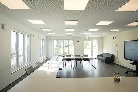 Ein heller Raum mit viel natürlichem Licht und Deckenleuchten sowie Tischen und Bestuhlung in U-Form.