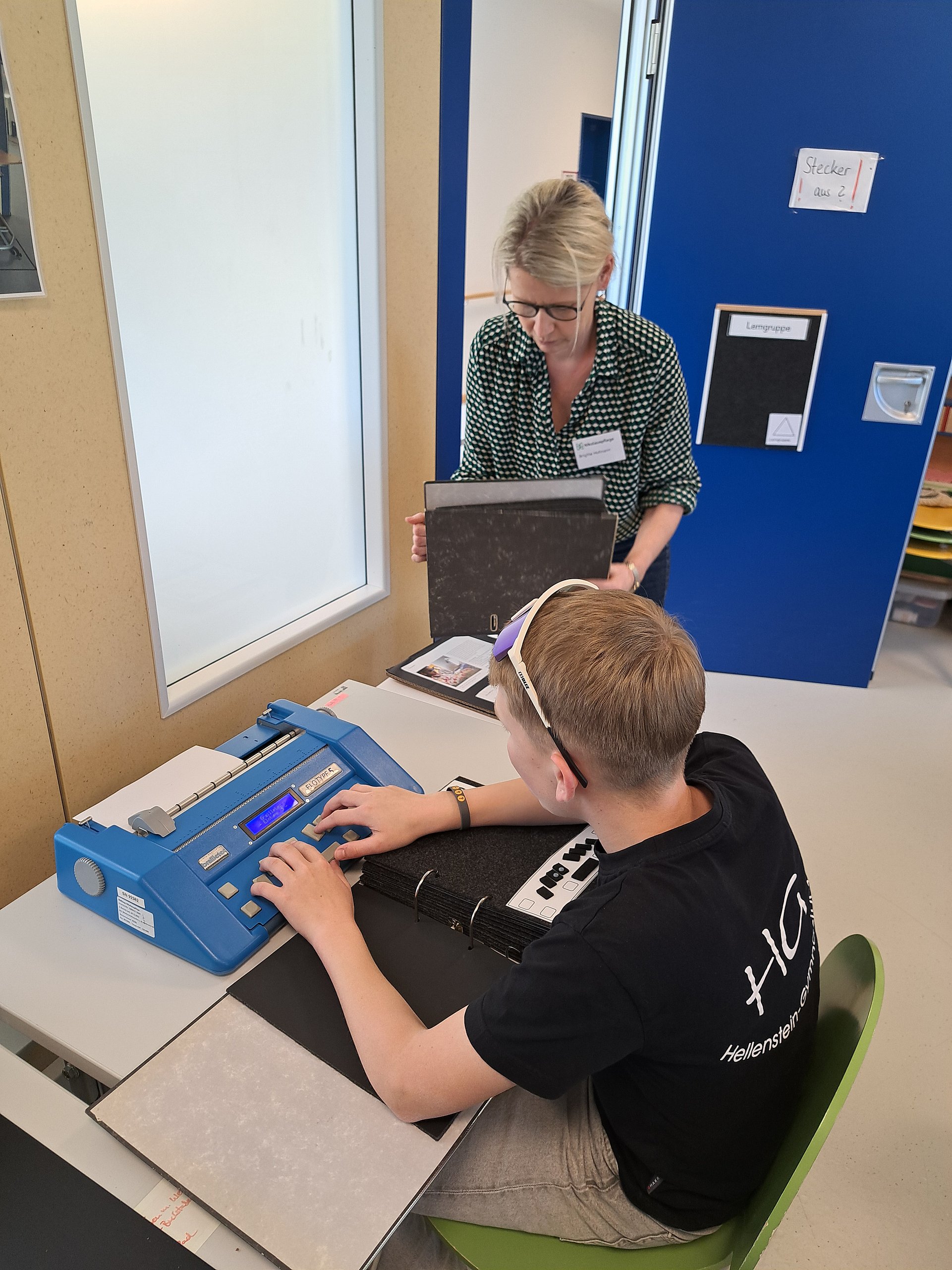Ein Schüler sitzt an einem Schreibtisch und schreibt auf einer Blindenschreibmaschine. Neben ihm steht eine Lehrerin, sie hält einen Ordner in der Hand