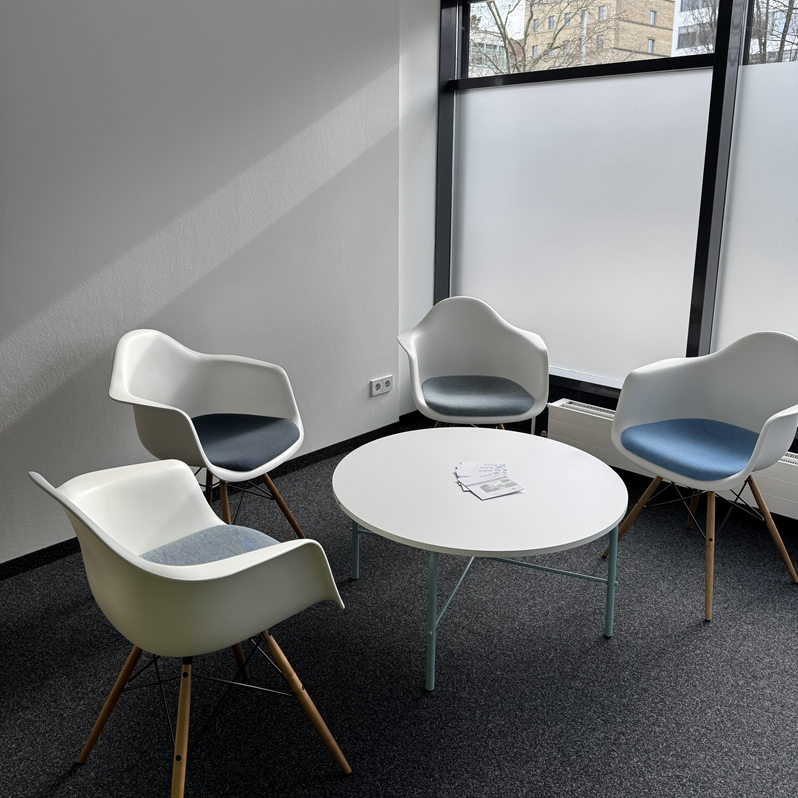 In einem hellen Raum mit grauem Teppichboden steht ein weißer runder Tisch. Darum sind vier weiße Stühle verteilt.