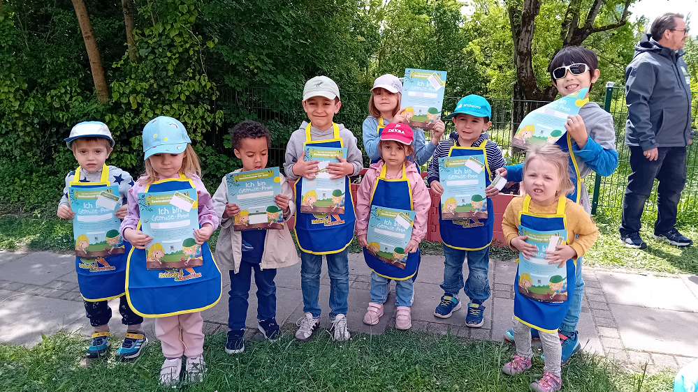 Neun Kinder stehen nebeneinander und lachen in die Kamera. Sie tragen gelb-blaue Schürzen, Mützen und halten eine Urkunde vor sich.