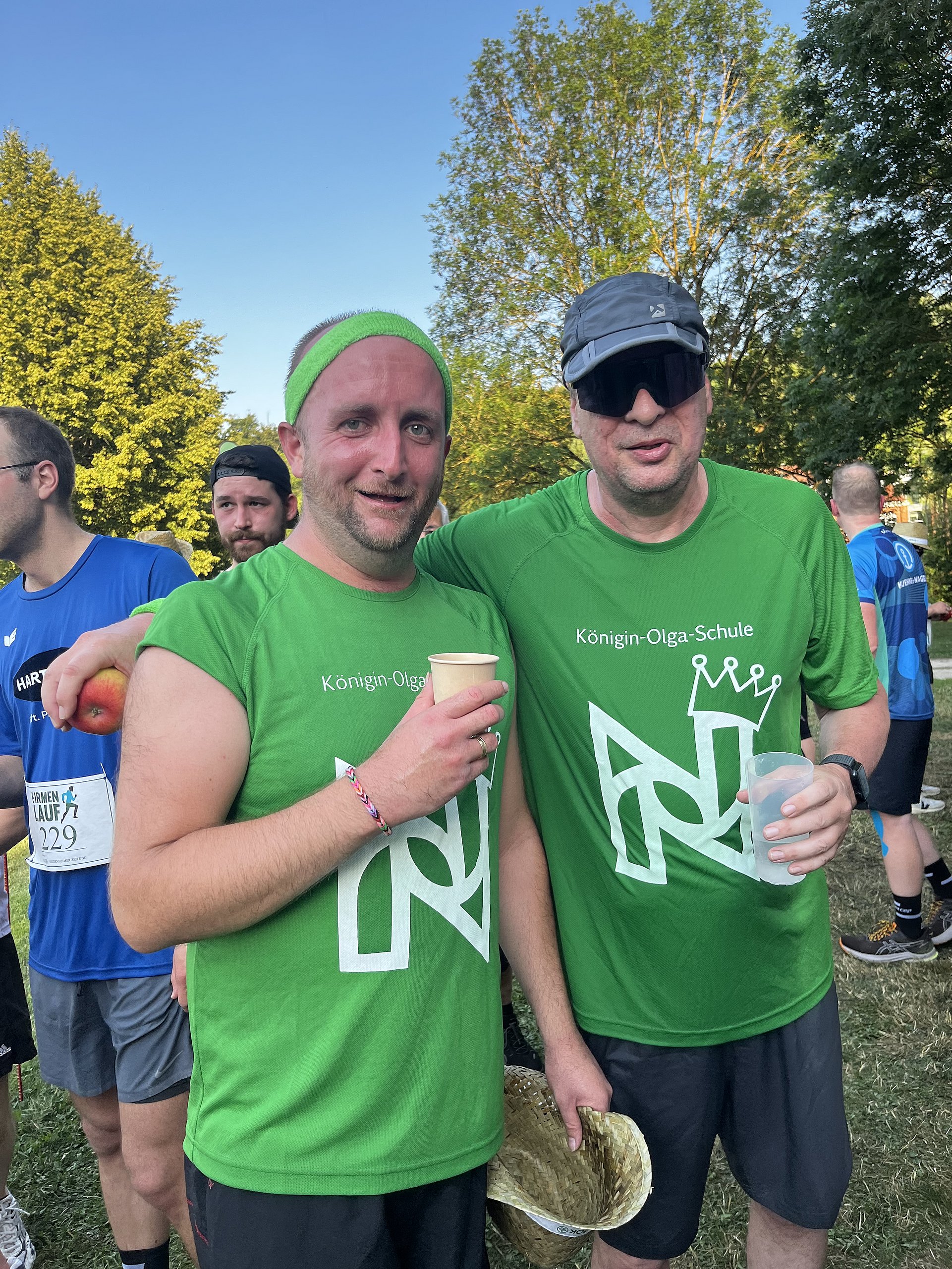 Zwei Männer in leuchtend grünen Laufshirts nach einem Laufveranstaltung.   Beide stehen dicht nebeneinander im Freien auf einer Wiese, im Hintergrund sind weitere Läufer, Bäume und Abendlicht.