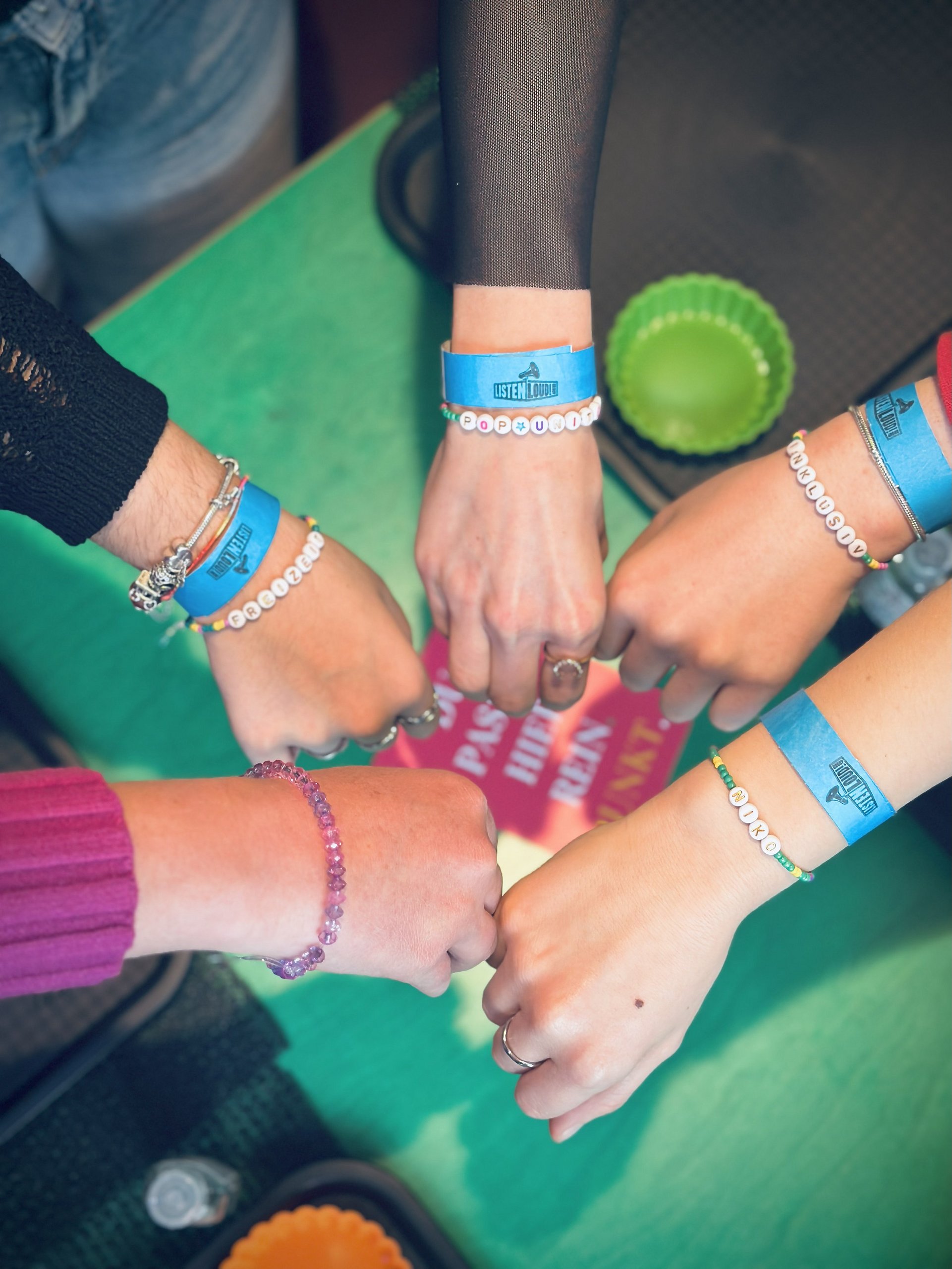 Fünf, zu Fäusten geballte Hände treffen sich in der Mitte des Bildes. an jedem Handgelenk hängen Perlenarmbänder