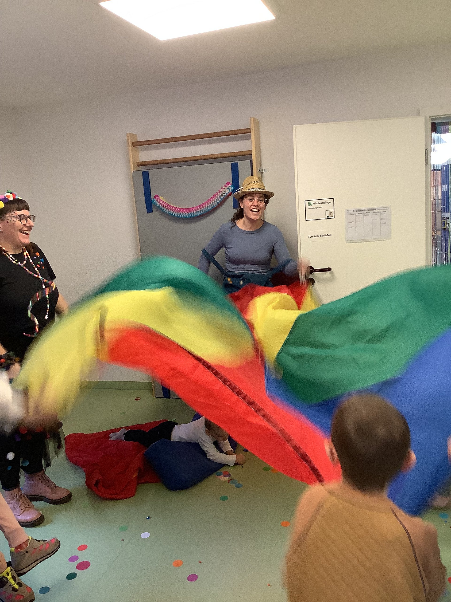 Betreuerinnen und Kinder stehen in einem Kreis und halten ein buntes Tuch, dass sie auf uns ab bewegen. Auf dem Boden liegt Konfetti