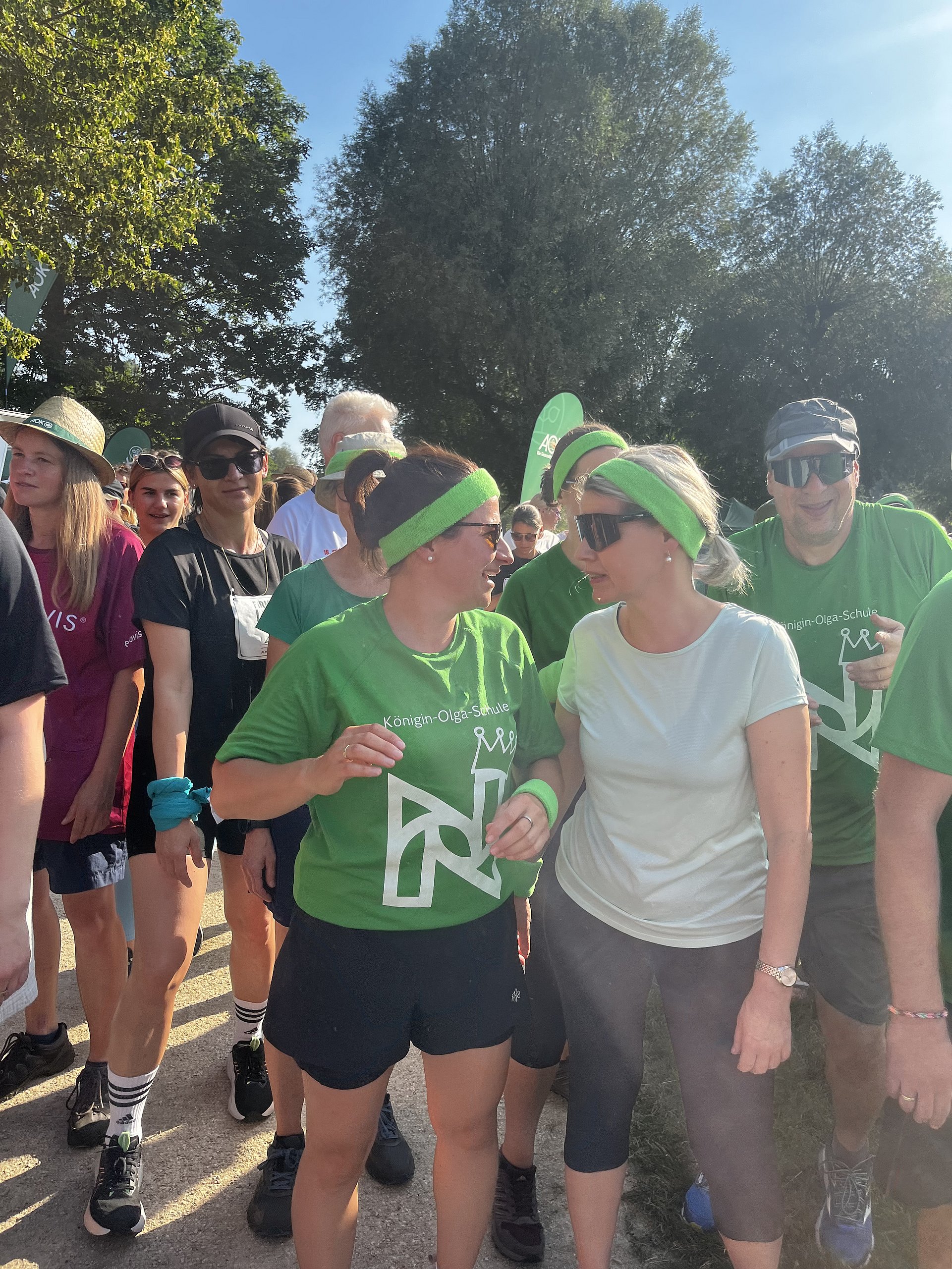 Eine Gruppe von Menschen bei einer Lauf- oder Wanderveranstaltung im Freien.   Im Vordergrund stehen zwei Frauen nebeneinander und unterhalten sich lächelnd. Die Frau links trägt ein grünes Stirnband, ein grünes T‑Shirt mit weißer Aufschrift „Königin-Olga-Schule“ und einem stilisierten Logo darauf, dazu dunkle Shorts und Sportschuhe. Die Frau rechts trägt ein helles T‑Shirt, eine dunkle dreiviertellange Sporthose, Sonnenbrille und ein hellgrünes Stirnband, ihre Haare sind zum Zopf gebunden.  