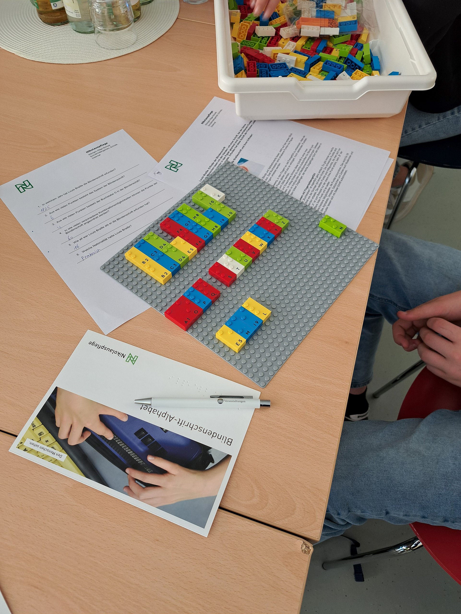 Auf einem Tisch liegen verschiedene Informationsbroschüren und eine Platte mit bunten Lego Braille Bausteine