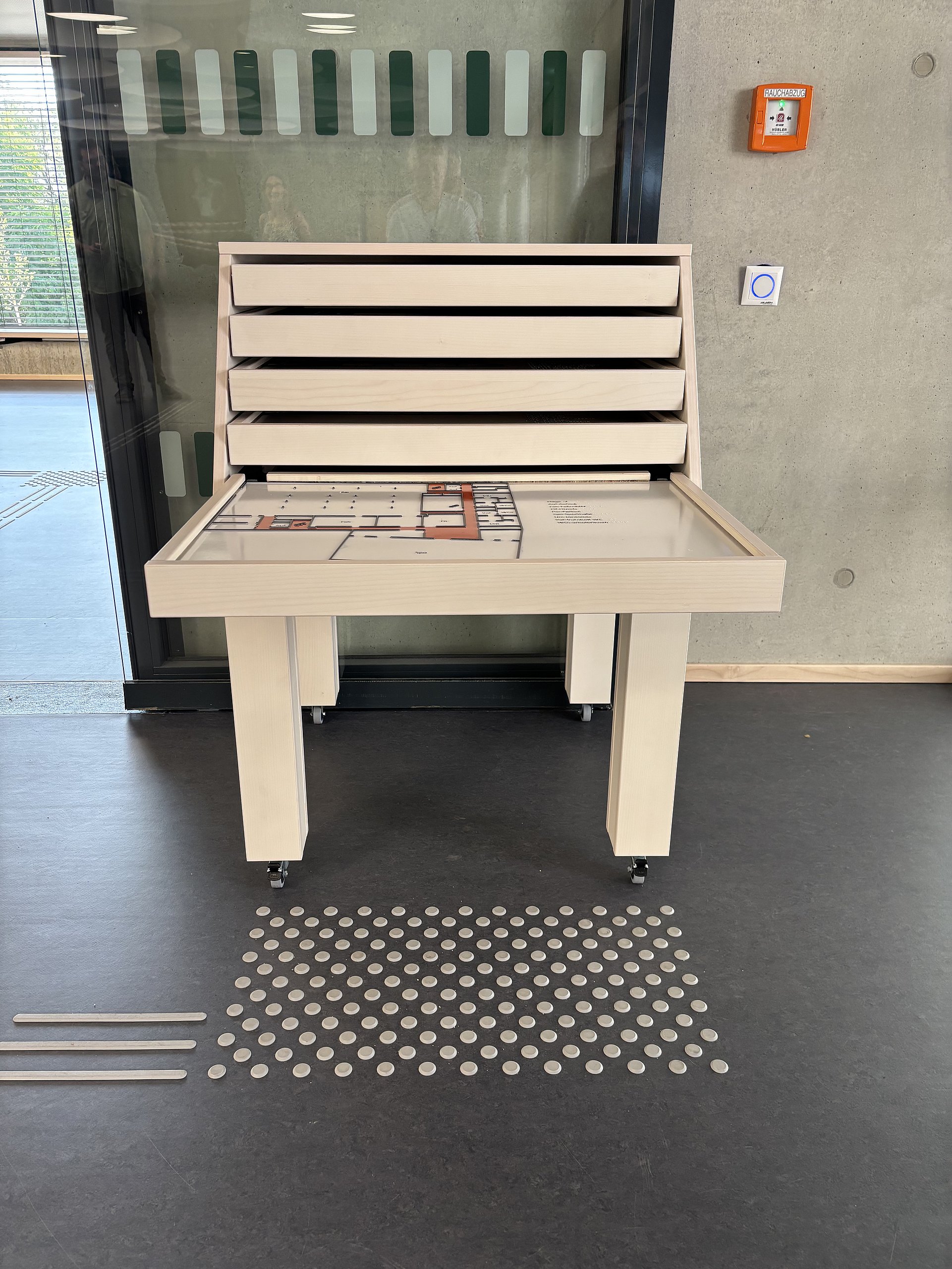 Ein hölzerner Informationstisch mit erhöhten Elementen steht auf einem dunklen Boden.   Auf dem Tisch liegt eine Art Plan oder Karte mit einem markierten Bereich, vermutlich ein Gebäudegrundriss; darauf sind verschiedene Rechtecke und Wege in Weiß, Grau und Orange zu erkennen.   Über der Tischfläche befindet sich ein senkrechter Aufbau mit mehreren waagerechten Leisten; sie wirken wie flache Schubladen oder Tafeln, die leicht nach vorne geneigt sind.  