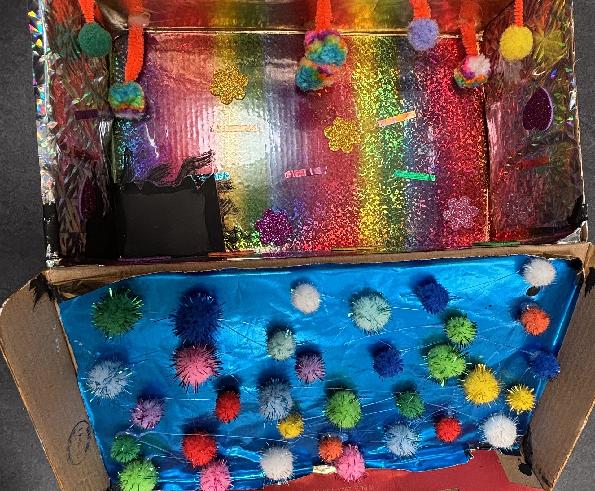 Ein aufgeklappter bunt gestalteter Pappkarton. Obere hälfte un glitzernden Regenbogenfarben, die untere in Blau und vielen bunten Filzkugeln