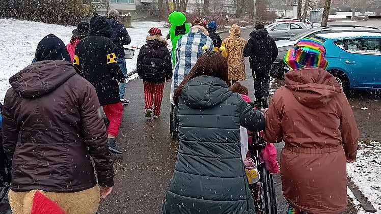 Mehrere Menschen in bunten Kostümen und mit Jacke spazieren eine Straße entlang.