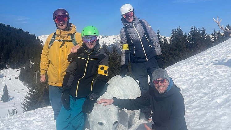Vier Personen, einer davon trägt eine Blindenarmbinde, stehen um eine Eisbärstatue herum, die an der Piste steht. Sie tragen alle Skiausrüstung und Helme.