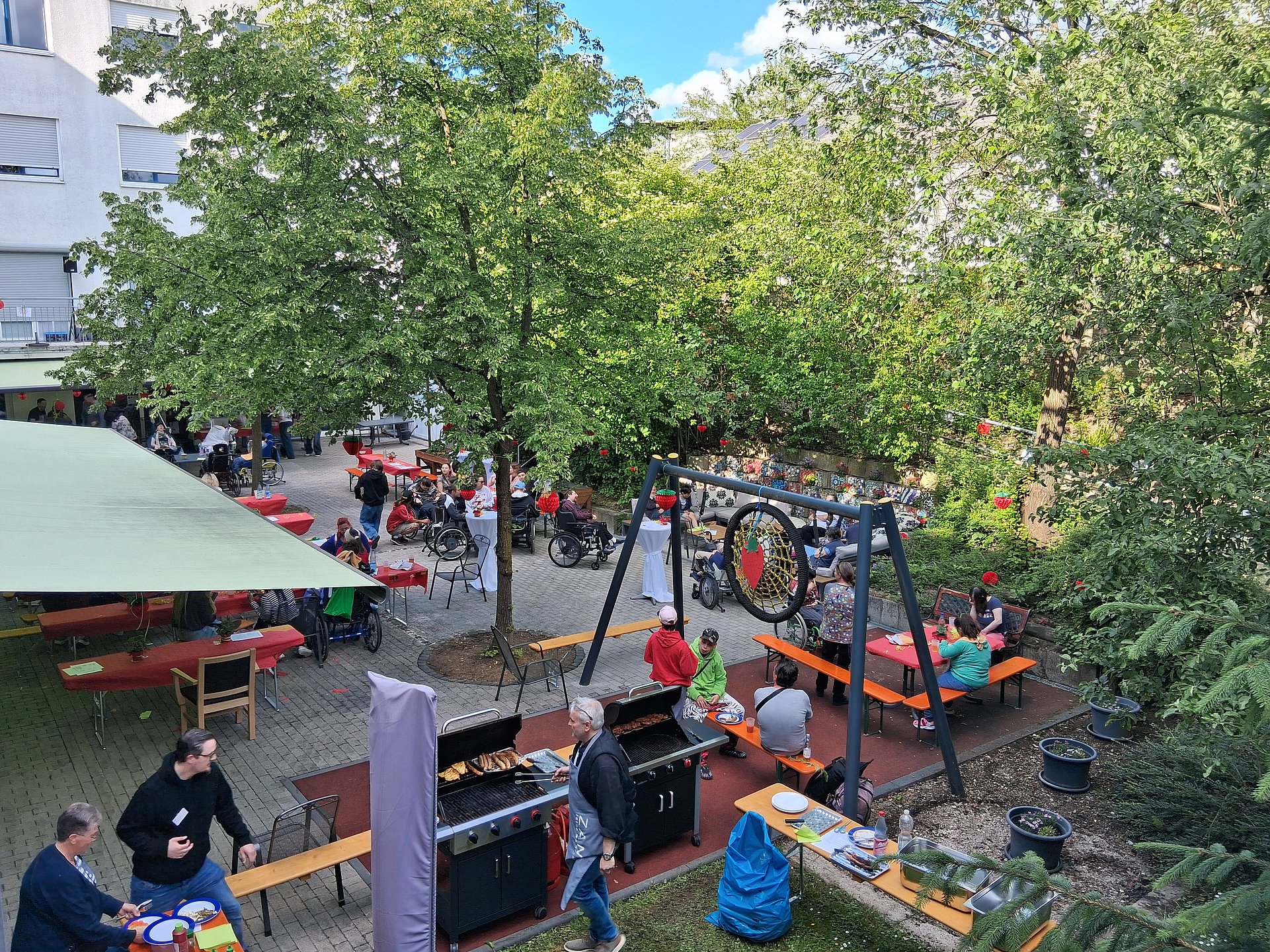 Der Außenbereich des Pau und Charlotte Kniese Hauses. Es sind Biertische und Bierbänke aufgestellt. Die Bäume haben grüne Blätter, der Himmel ist blau. Man sieht viele Menschen. 