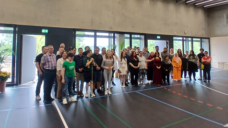 Zahlreiche Jugendliche und Erwachsene stehen in einer Sporthalle vor einer Fensterfront in drei Reihen hintereinander und blicken rechts an der Kamera vorbei. Ein junger Mann hält einen Blindenstock in der Hand.