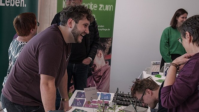 Ein junger Mann steht hinter einem großen Tisch, auf dem Flyer und Materialien ausgebreitet sind. Hinter ihm stehen zwei große Rollups der Nikolauspflege.  Er lächelt und unterhält sich mit Personen, auf der anderen Seite des Tisches.