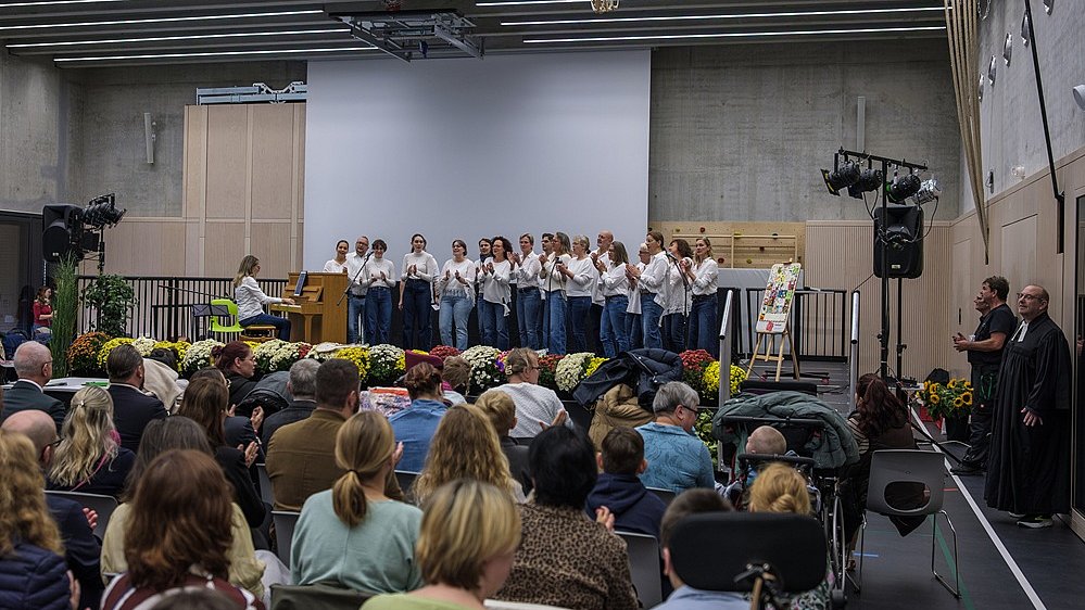 In der Sporthalle sitzen zahlreiche Menschen auf Stühlen und blicken nach vorne zur Bühne. Darauf steht eine Gruppe Personen, die alle dunkle Hosen und weiße Oberteile tragen. Neben ihnen spielt eine Frau an einem Klavier.