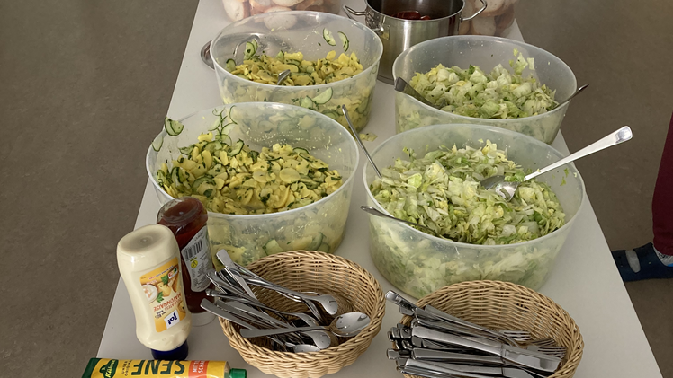 Auf einem Tisch ist ein Holztablett, darauf mehrere Schüsseln mit Kartoffelsalat und Brotscheiben, zwei Körbe mit Besteck, eine Tube Mayonnaise und Ketchup und eine Tube Senf.