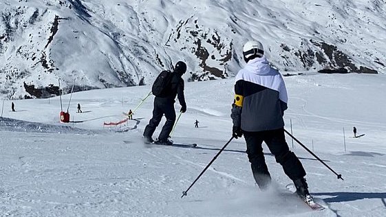 Zwei Menschen, einer davon trägt eine Blindenarmbinde, fahren auf Ski die Piste hinunter.