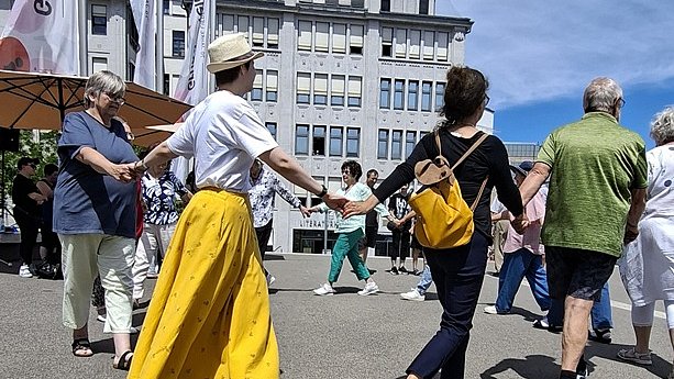 Mehrere Menschen, die sich an den Händen halten und im Kreis tanzen, auf einem Platz im Freien. Im Vordergrund links trägt eine Person mit heller Haut einen langen, leuchtend gelben Rock, ein weißes T Shirt, flache schwarze Sandalen und einen beigefarbenen Strohhut. Daneben tanzt eine Person mit dunkler Oberbekleidung und dunkler Dreiviertelhose, helle Haut, barfuß, mit einem orangefarbenen Rucksack auf dem Rücken. Rechts davon sind weitere tanzende Menschen mit heller Haut zu erkennen; sie tragen überwiegend Sommerkleidung wie T Shirts, Shorts oder leichte Hosen in Grün, Blau und Beige. Im Hintergrund stehen noch mehr Personen und mehrere hohe Fahnenmasten mit weißen Fahnen und roten Elementen. Dahinter befindet sich eine graue mehrstöckige Häuserzeile mit vielen Fenstern und roten Dächern. Der Himmel ist blau mit leichten Wolken, das Licht wirkt sonnig und kräftig.