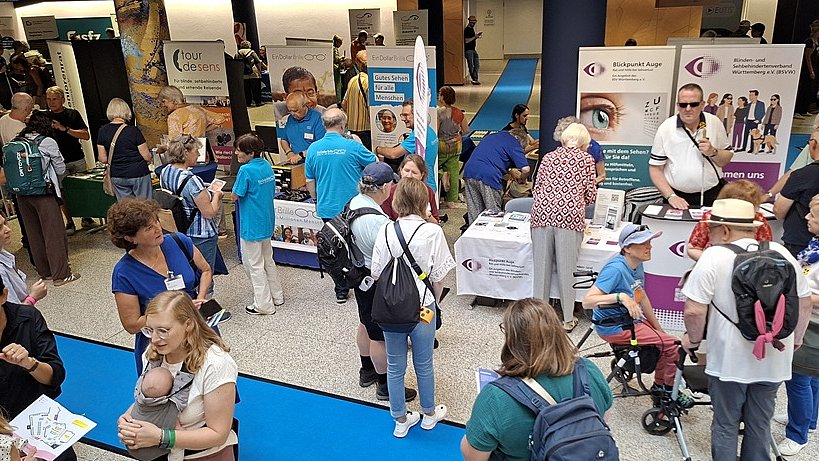Großer Veranstaltungssaal mit vielen Menschen auf einer Messe: Mehrere Stände stehen dicht nebeneinander auf einem hellen Holzfußboden. Vorne links befindet sich ein Metallständer mit mehreren Reihen Flyern. Dahinter steht ein gelber, mit Stoff bedeckter Tisch mit Infomaterial. Darüber hängt ein großes gelbes Banner mit dem Schriftzug „Reinecker“ und dem Foto eines älteren Mannes und einer älteren Frau mit Brille, beide lächeln. Viele Personen stehen oder gehen durch die Gänge, einige mit Rollatoren, andere im Gespräch an den Ständen. Die Menschen tragen überwiegend Freizeitkleidung wie T Shirts, kurze Hosen oder Sommerkleider. Der Raum ist hoch und modern mit grauen Wänden und einer geschwungenen Decke mit vielen kleinen Einbauleuchten. Rechts oben ist eine Empore zu erkennen. An der Decke hängen zahlreiche Scheinwerfer und technische Elemente.