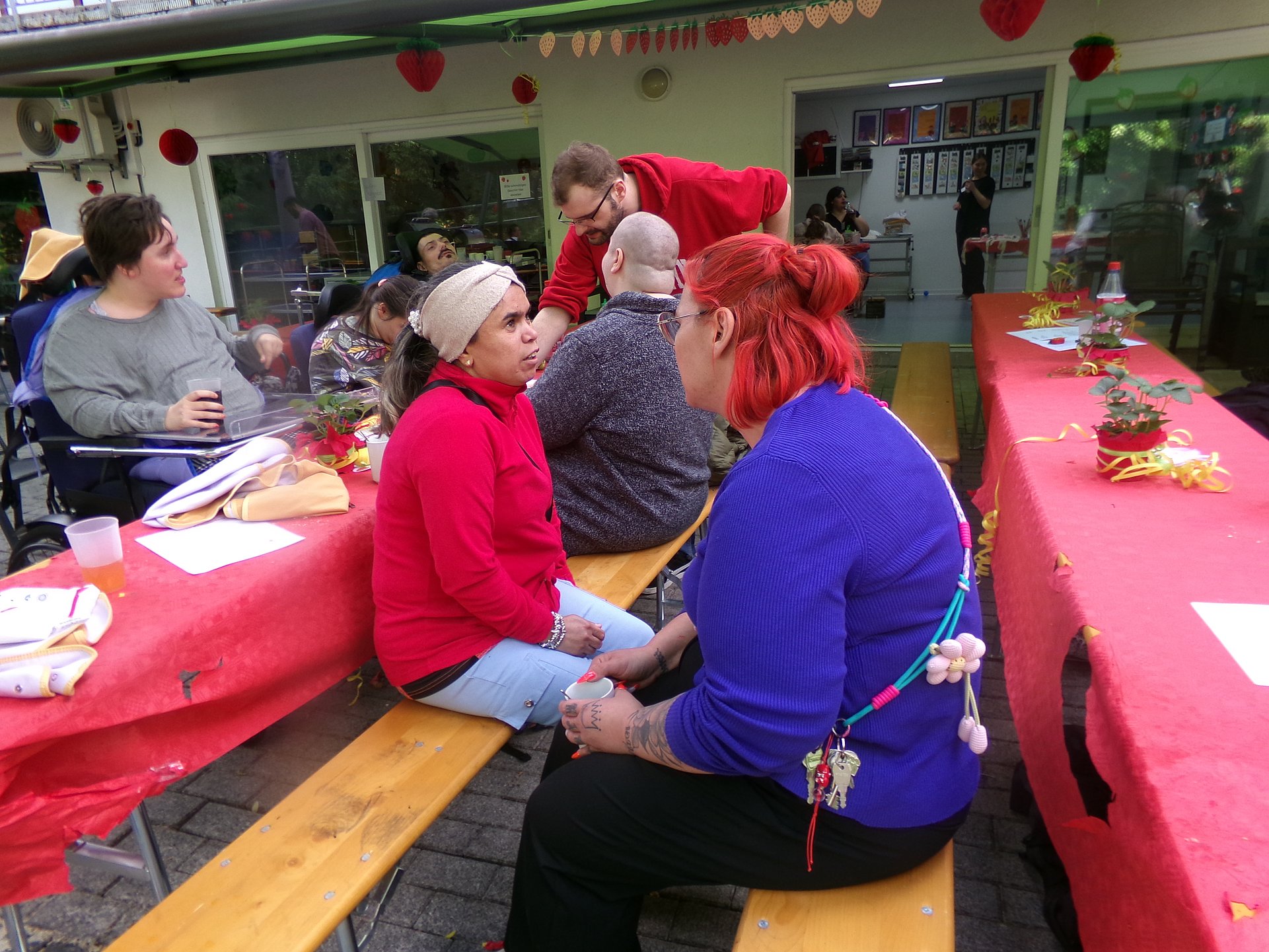 Zwei Frauen sitzen auf zwei Bierbänken gegenüber und unterhalten sich. Hinter ihnen stehen Stische mit roten Tischdecken. Die Tische sind gedeckt. Um sie herum Stehen Menschen