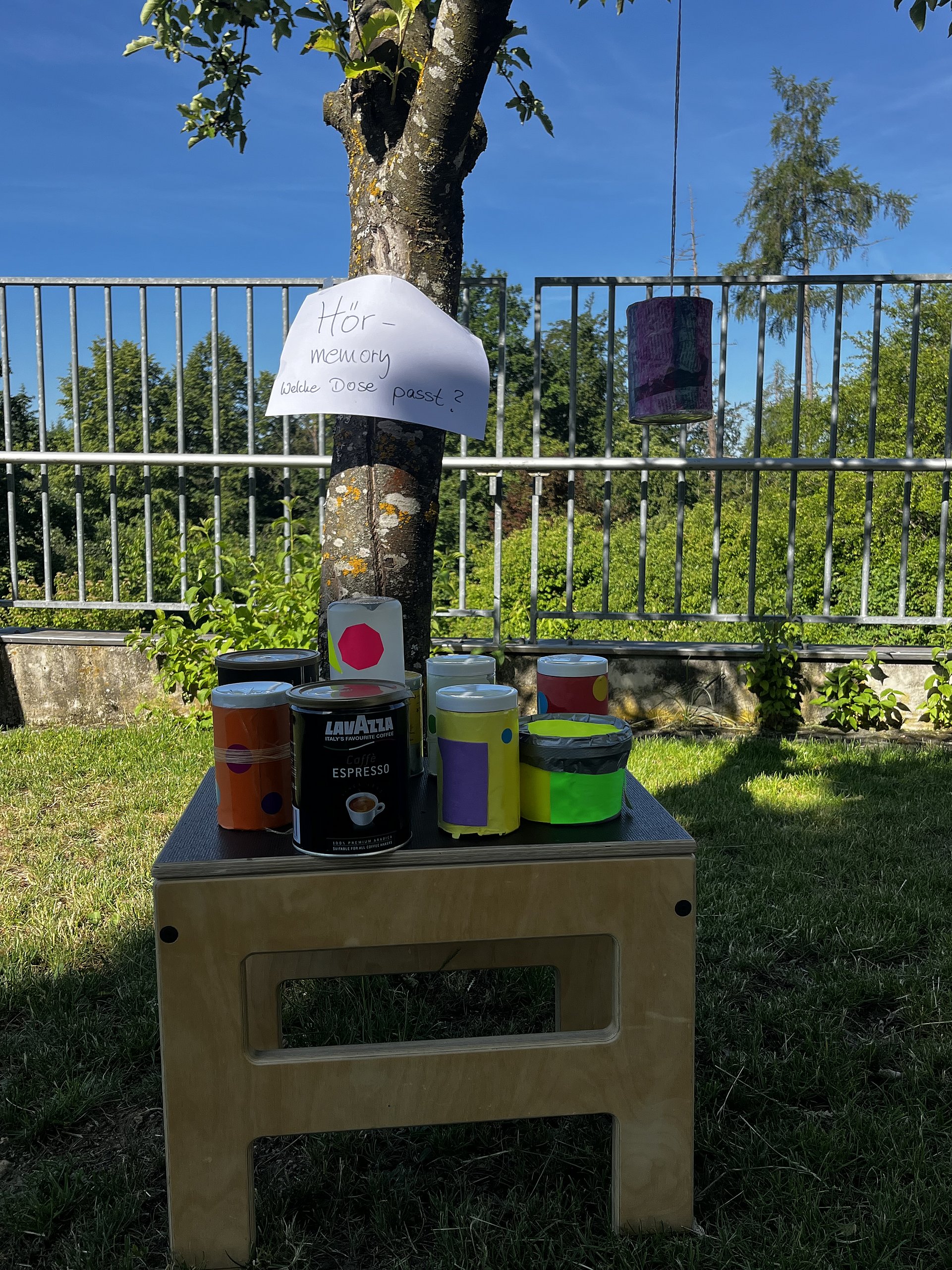 Ein kleiner Holzhocker mit mehreren bunt beklebten Dosen darauf im Freien.   Auf dem Hocker stehen von links nach rechts: Hinter dem Hocker steht ein Baum. An dem Baum ist ein weißes Blatt Papier befestigt, darauf steht handgeschrieben:„Hör – Memory Welche Dose passt?“  Rechts vom Baum hängt an einer Schnur eine lila Dose oder ein lila beklebter Zylinder in ungefähr Augenhöhe, offenbar als Teil des Spiels.