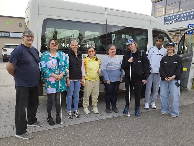 Ein Gruppenfoto vor türkisfarbenem Hintergrund: Acht Personen stehen draußen nebeneinander vor einem grauen Van. Einige Personen halten weiße Langstöcke in der Hand. Im Hintergrund ist ein Gebäudeteil zu erkennen. Im unteren Bild ist ein weißer Kasten mit schwarzem Text: „„Klientinnen und Klienten beim Schnupperkurs zur Selbstbehauptung und Selbstverteidigung“.