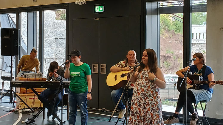 In einer Sporthalle vor einer Fensterfront stehen und sitzen sechs Personen. Sie spielen gemeinsam in einer Band, es gibt ein Keyboard, zwei an der Gitarre und zwei am Mikrofon.