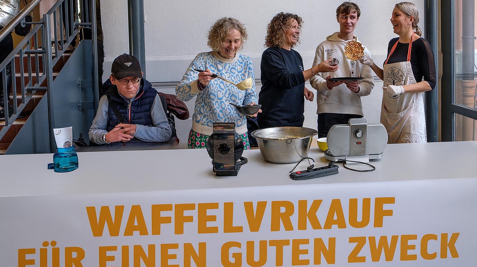 Mitarbeitende und Teilnehmervertretende stehen hinter einem langen Tisch. Dieser ist mit einer weißen Tischdecke gedeckt. Darauf stehen Schüsseln und ein Waffeleisen. Eine Mitarbeiterin schöpft Teig mit einer Schöpfkelle aus einer Schüssel. 