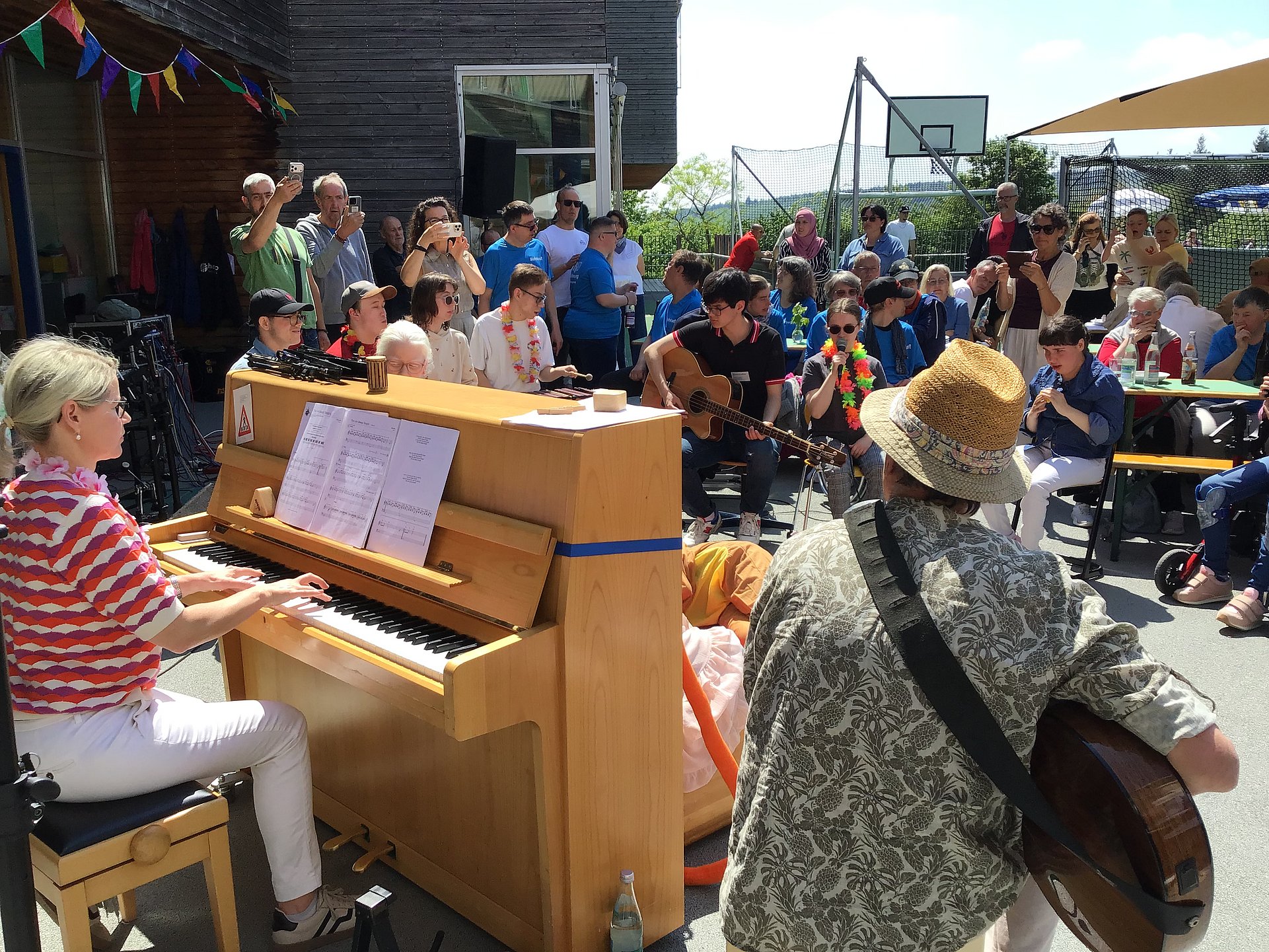 Auf dem Foto sitzt links eine Person mit blonden Haaren, rot‑weiß gestreiftem Oberteil und weißer Hose an einem hellen Holzklavier oder -orgel und spielt. Vor ihr liegt ein aufgeklapptes Notenblatt auf dem Instrument. Rechts und im Hintergrund steht und sitzt eine größere Gruppe von Menschen in sommerlicher Kleidung, die der Musik zuhören; einige tragen bunte Accessoires wie Hüte oder Blumenketten. Im unteren Bildbereich ist eine Person mit Strohhut und gemustertem Hemd von hinten zu erkennen, die ein Saiteninstrument mit rundem Korpus umgehängt hat. Im Hintergrund des Fotos sind ein Basketballkorb, Tische und Bänke sowie bunte Wimpel zu erkennen