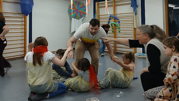 Ein Mann steht in der Mitte eines Raums um ihn herum sind drei Kinder und eine junge Frau, sie tragen gelbe Müllsäcke um den Körper und zerren an dem Mann in der Mitte.