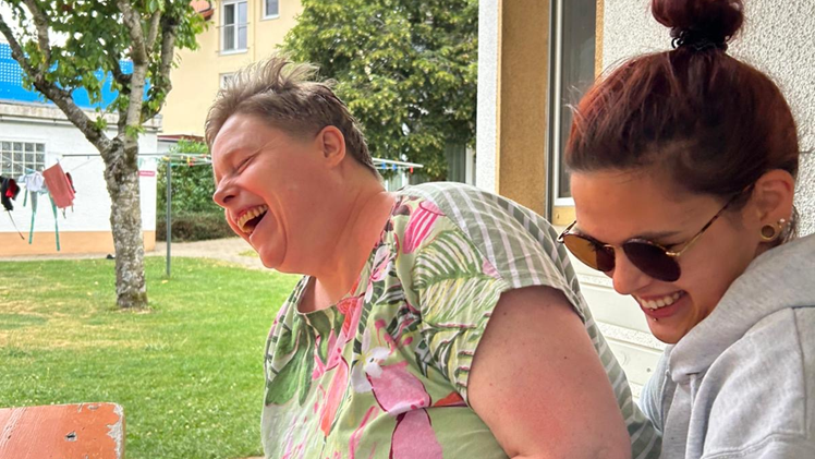 ine Frau mit grün-pink-geblümtem T-Shirt und kurzen Haaren sitzt vor einem Haus. Links neben ihr sitzt eine junge Frau in grauem Pullover, mit langen dunklen Haaren und schwarzer Sonnenbrille. Beide Frauen lachen herzlich. Im Hintergrund sind mehrere Häuser, eine Wiese und ein Baum.