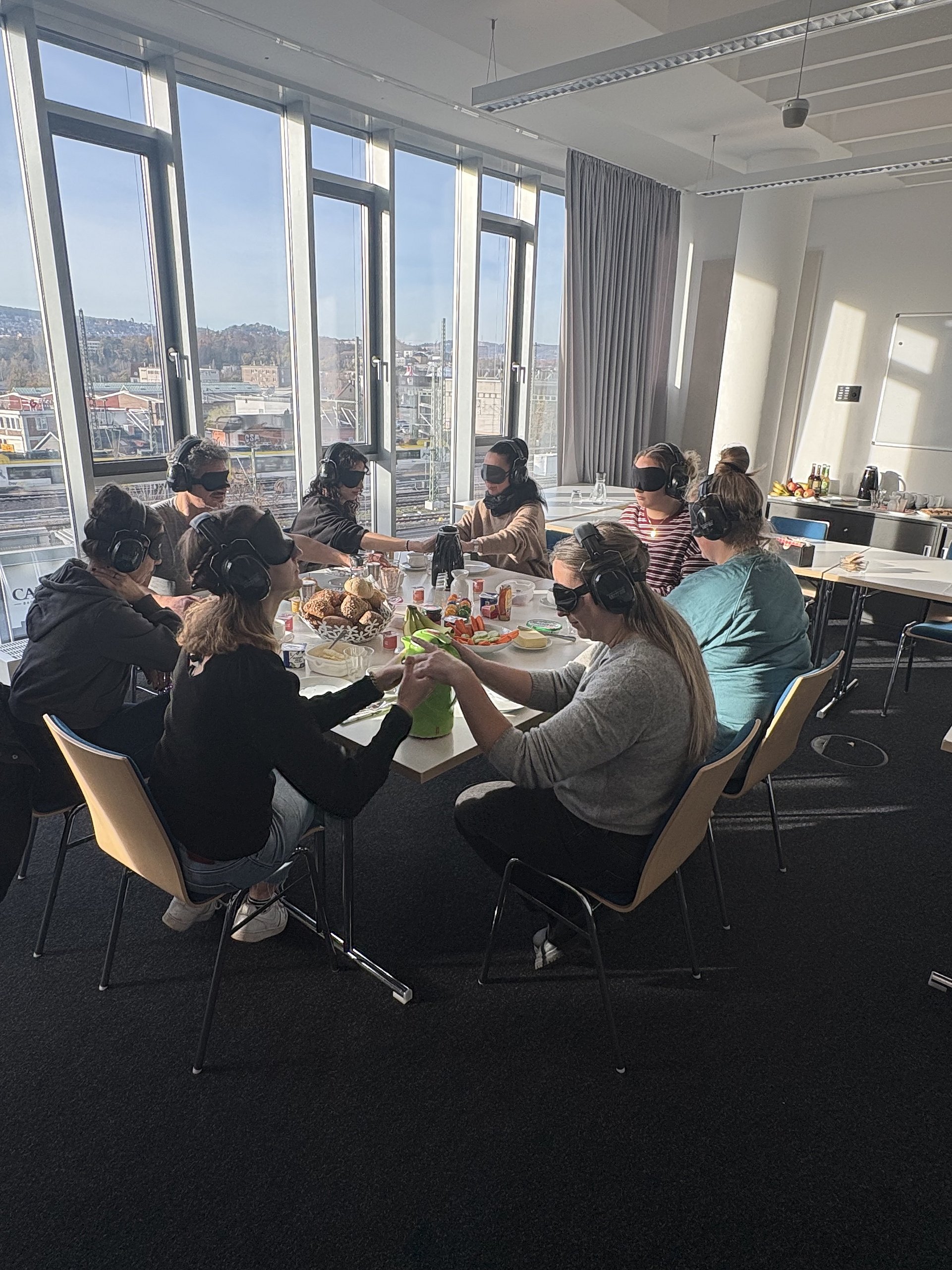 Eine Gruppe von Mitarbeitenden sitzt in einem Konferenzraum gemeinsam an einem Tisch. Alle tragen Augenbinden und Kopfhörer. Sie essen und trinken