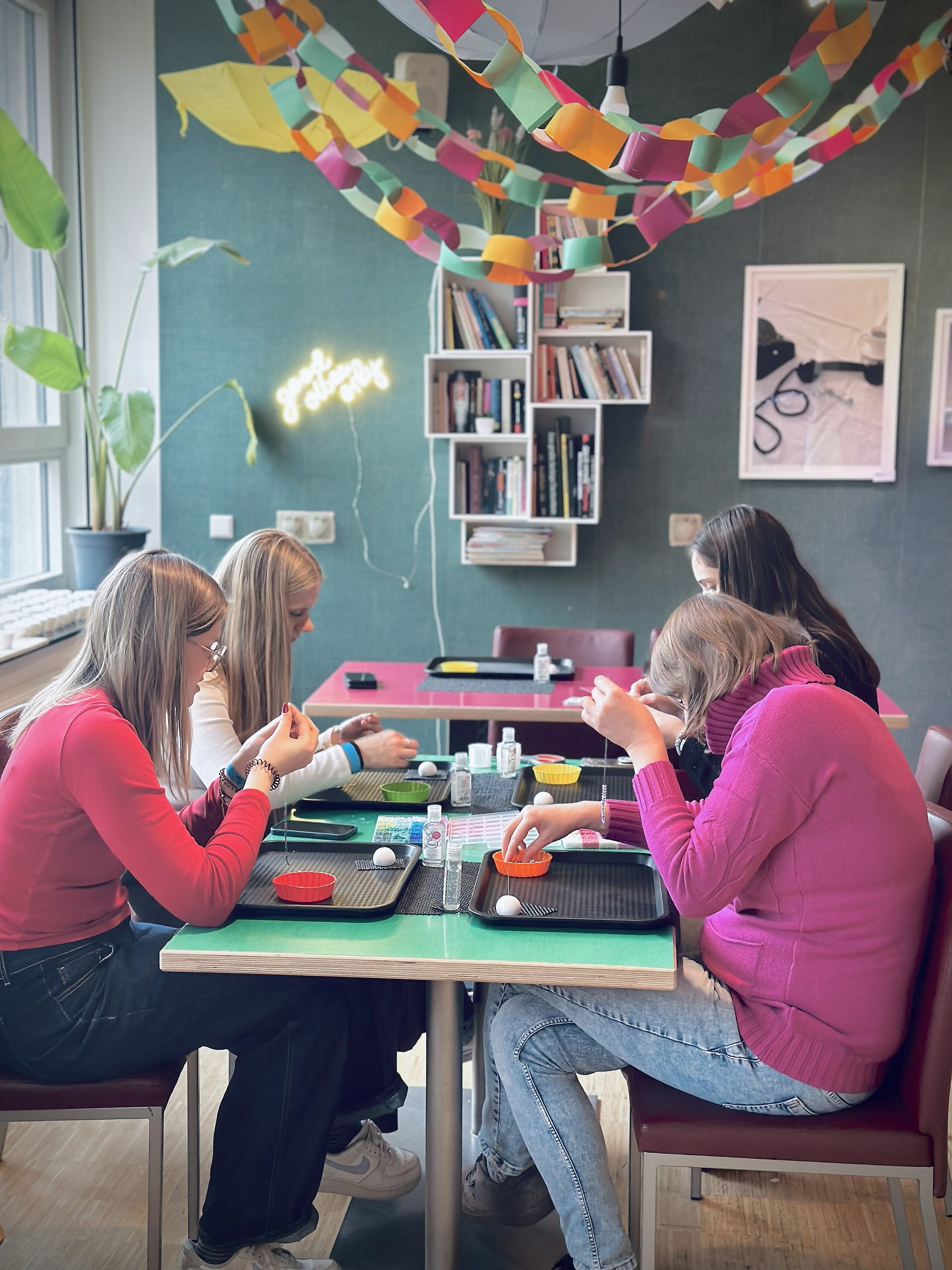 Vier junge Frauen sitzen gemeinsam an einem Tisch in einem bunt dekorierten Raum. Sie fädeln Perlen an Schnüre zu Armbänder