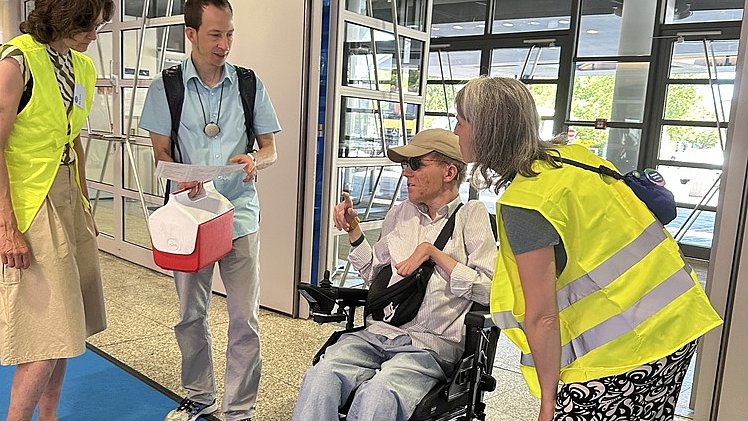 Vier Personen in einem Eingangsbereich auf einem blauen Teppich. In der Bildmitte sitzt eine Person mit heller Haut in einem elektrischen Rollstuhl, trägt eine hellgraue Hose, helle Turnschuhe, ein helles Oberteil und eine beigefarbene Kappe. Vor der Person hängt eine schwarze Umhängetasche, in der rechten Hand hält sie ein Blatt Papier. Links davon steht eine Frau mit heller Haut in einem gelben Warnüberwurf über einem ärmellosen Oberteil und einem knielangen beigen Rock, an den Füßen Sandalen. Neben ihr steht ein Mann mit heller Haut in einem hellblauen Kurzarmhemd und grauer Hose, mit Rucksackgurten über den Schultern und einem Ausweisband um den Hals; er hält eine weiße Mappe und eine rot weiße Box in den Händen. Rechts vom Rollstuhl beugt sich eine weitere Frau mit heller Haut vor; sie trägt ebenfalls eine gelbe Warnweste, ein schwarzes T Shirt und weite schwarz weiß gemusterte Hose mit flachen Sandalen. Alle scheinen miteinander zu sprechen. Im Hintergrund befinden sich große Glas- und Metalltüren eines modernen Gebäudeeingangs mit hellem Boden.