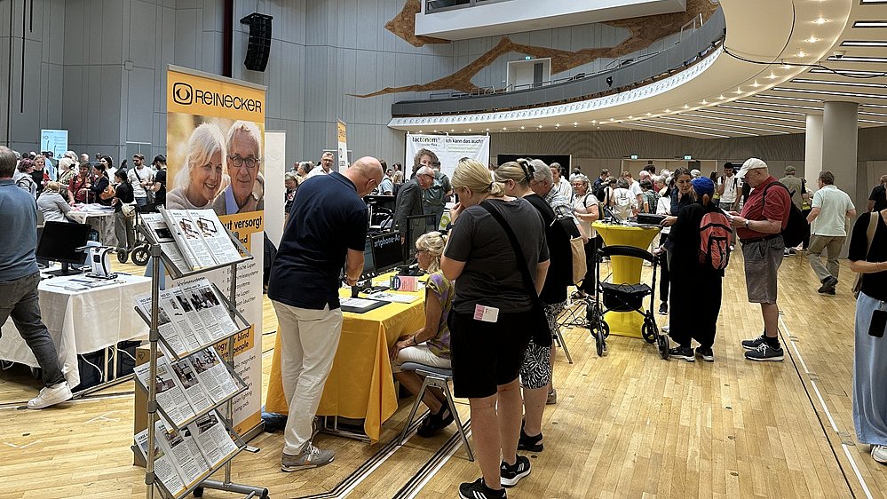 Messestand-Szene auf einer Messe: Mehrere Stände mit Roll Up Bannern und Tischen sind eng nebeneinander aufgebaut, davor stehen und gehen viele Menschen unterschiedlichen Alters, einige mit Rucksäcken, Kinderwagen oder Hilfsmitteln wie einem Rollator. Viele Personen tragen Namensschilder, einige haben türkisfarbene T Shirts an. Vorne links sitzt eine Frau mit hellblonden Haaren und heller Haut auf einer Bank, sie hält ein kleines Kind auf dem Arm. Daneben verläuft ein blauer Läufer auf dem Boden. Weiter hinten stehen Gruppen von Menschen an Tischen, auf denen Prospekte, Flyer und Broschüren ausgelegt sind. Auf einem Tisch ist eine rot weiße Tischdecke mit einem Muster zu erkennen. In der oberen Bildhälfte sind weitere Besucherinnen und Besucher vor einem Bereich mit holzfarbenen Wänden und warmem Licht, dort hängen große Banner, die auf weitere Angebote hinweisen. Insgesamt vermittelt das Bild eine dichte, lebhafte Atmosphäre mit vielen Gesprächen und Informationsaustausch.