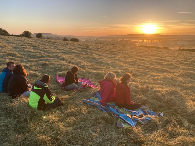 Eine Gruppe von Menschen sitzen gemeinsam auf Decken auf einer Wiese auf einem Hügel. Sie betrachten den Sonnenaufgang am Horizont