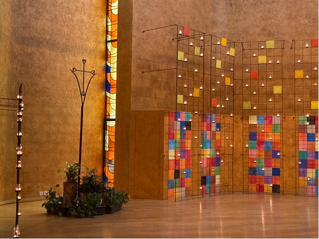 Innenansicht der Kirche des Männerordens in Taisé in Frankreich. Die Wände sind mit bunten Kacheln geschmückt. In Gestellen leuchten Kerzen. Durch bunte Fenstergläser fällt Tageslicht. Seitlich im Raum steht ein Ständer, darunter befinden sich einige Topfpflanzen.