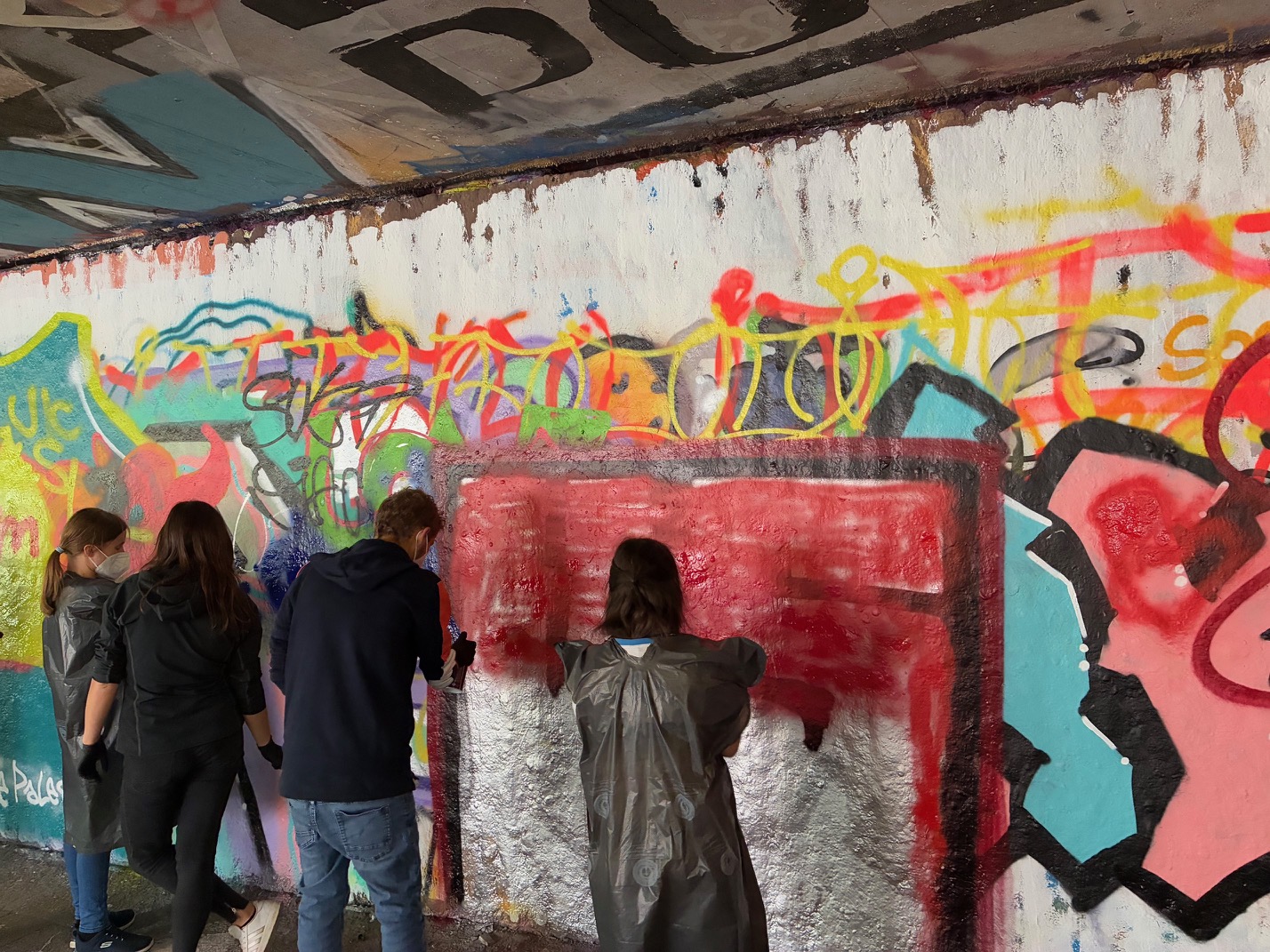 Vier Jugendliche stehen vor einer bunten Wand und sprühen mit Spraydosen gemeinsam an einem Graffitti