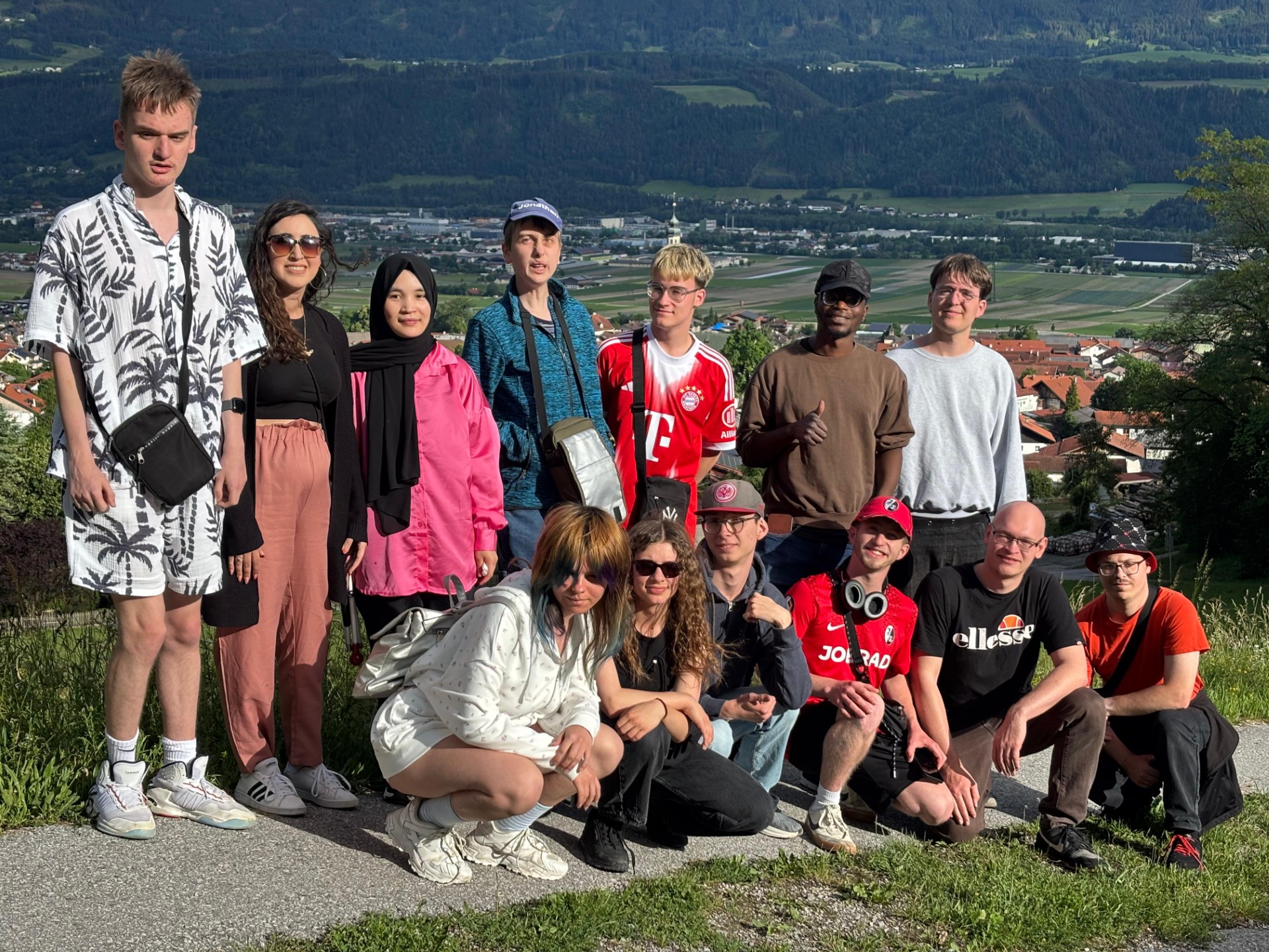 Eine größere Gruppe junger Menschen sitzt und steht im Freien vor einer Berglandschaft.   Im Vordergrund sitzt eine Reihe von Personen auf einem niedrigen Mäuerchen oder Steinrand. Alle wirken entspannt und posieren nah beieinander für ein Gruppenfoto. Im Hintergrund erstreckt sich ein weitläufiges Tal mit Feldern, einem Ort mit orangefarbenen Dächern und hohen grünen Bergen dahinter.
