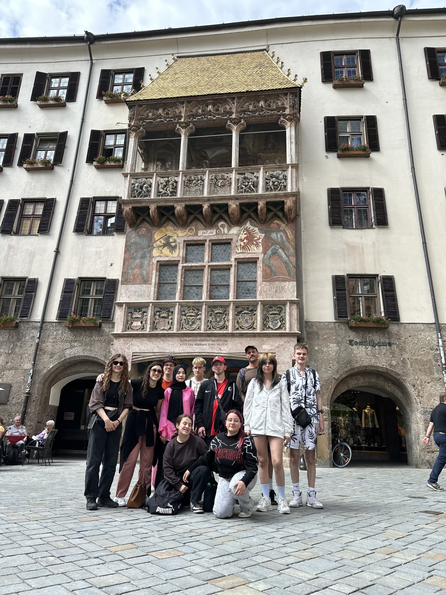 Eine Gruppe junger Menschen vor einer historischen Hausfassade mit reich verziertem Erker.   Vor dem Gebäude steht eine größere Gruppe, etwa zwölf Personen, die in zwei Reihen posieren: einige hocken vorne auf dem Pflaster, die anderen stehen dahinter. Die Kleidung ist modern und lässig – überwiegend dunkle Töne, dazu einzelne auffällige Farben wie ein leuchtend pinkes Outfit und eine weiße, langärmelige Kapuzenjacke mit kurzen Hosen. Mehrere Personen tragen Turnschuhe, einige haben Rucksäcke oder Umhängetaschen. Die Stimmung wirkt freundlich und wie bei einem Ausflugsfoto.