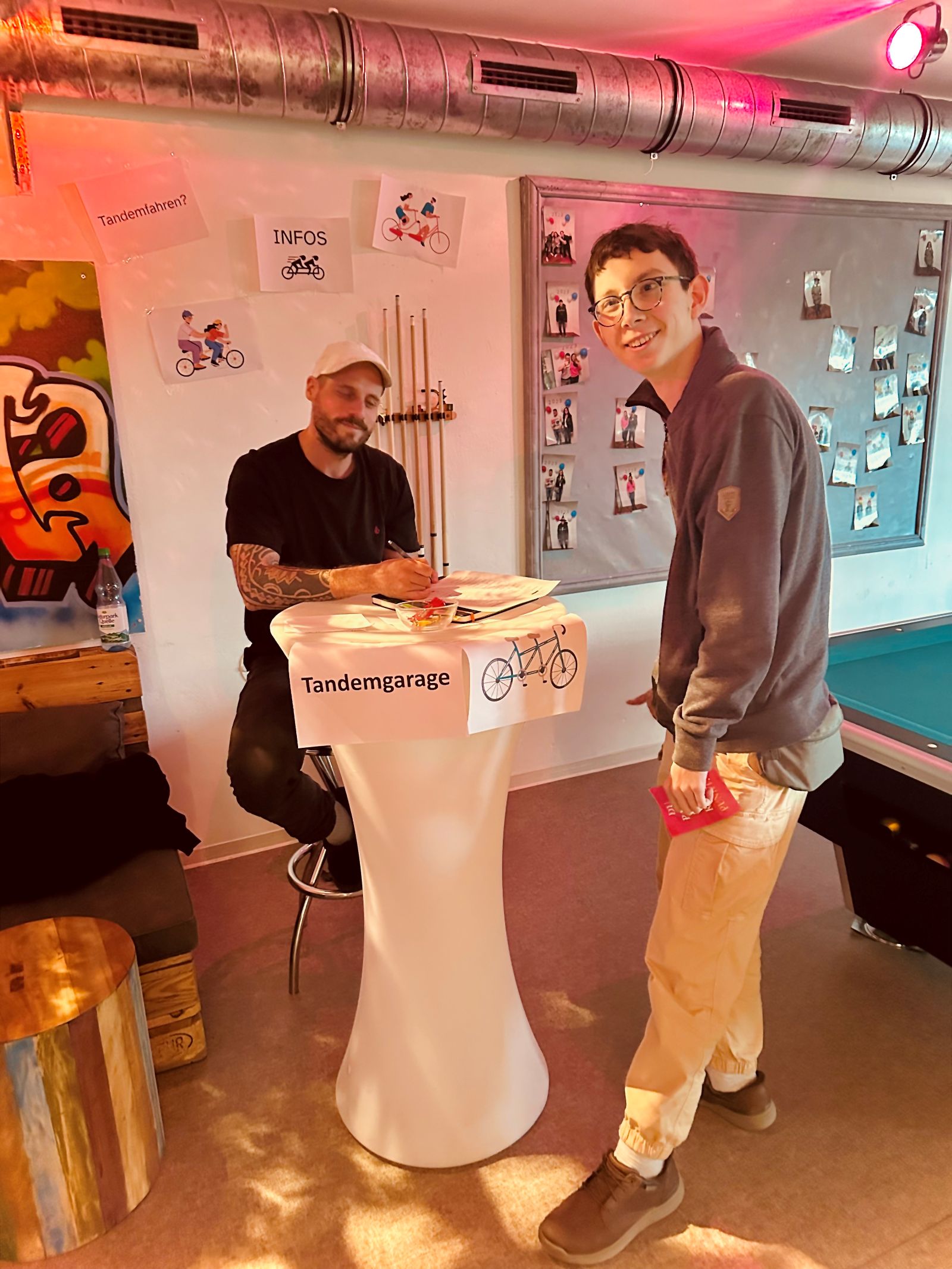 Ein junger Mann steht an einem Informationsstand. Dahinter steht noch ein Mann. Beide lächeln. An dem Stand wird über Tandemfahrten informiert