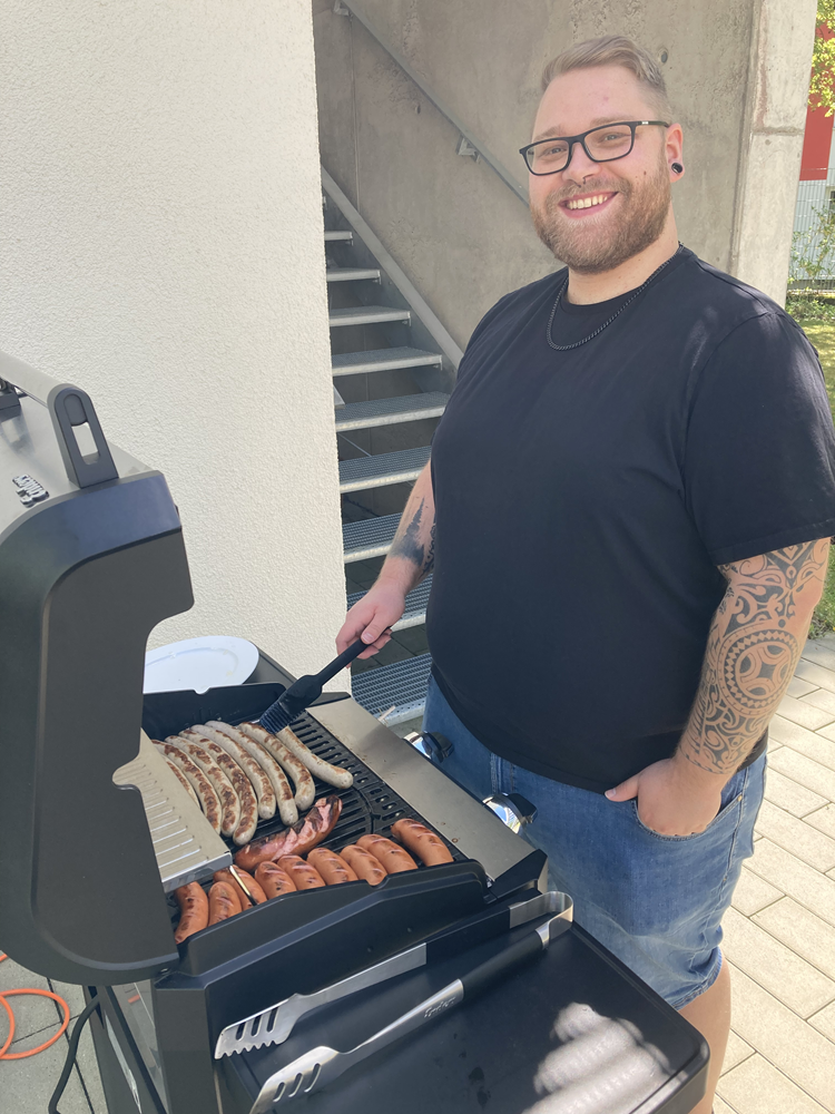 Ein Mann in Jeans, schwarzem T-Shirt, schwarzer Brille, kurzen Haaren und Bart steht an einem Grill und lacht in die Kamera. Auf dem Grill liegen zahlreiche rote und weiße Würstchen.